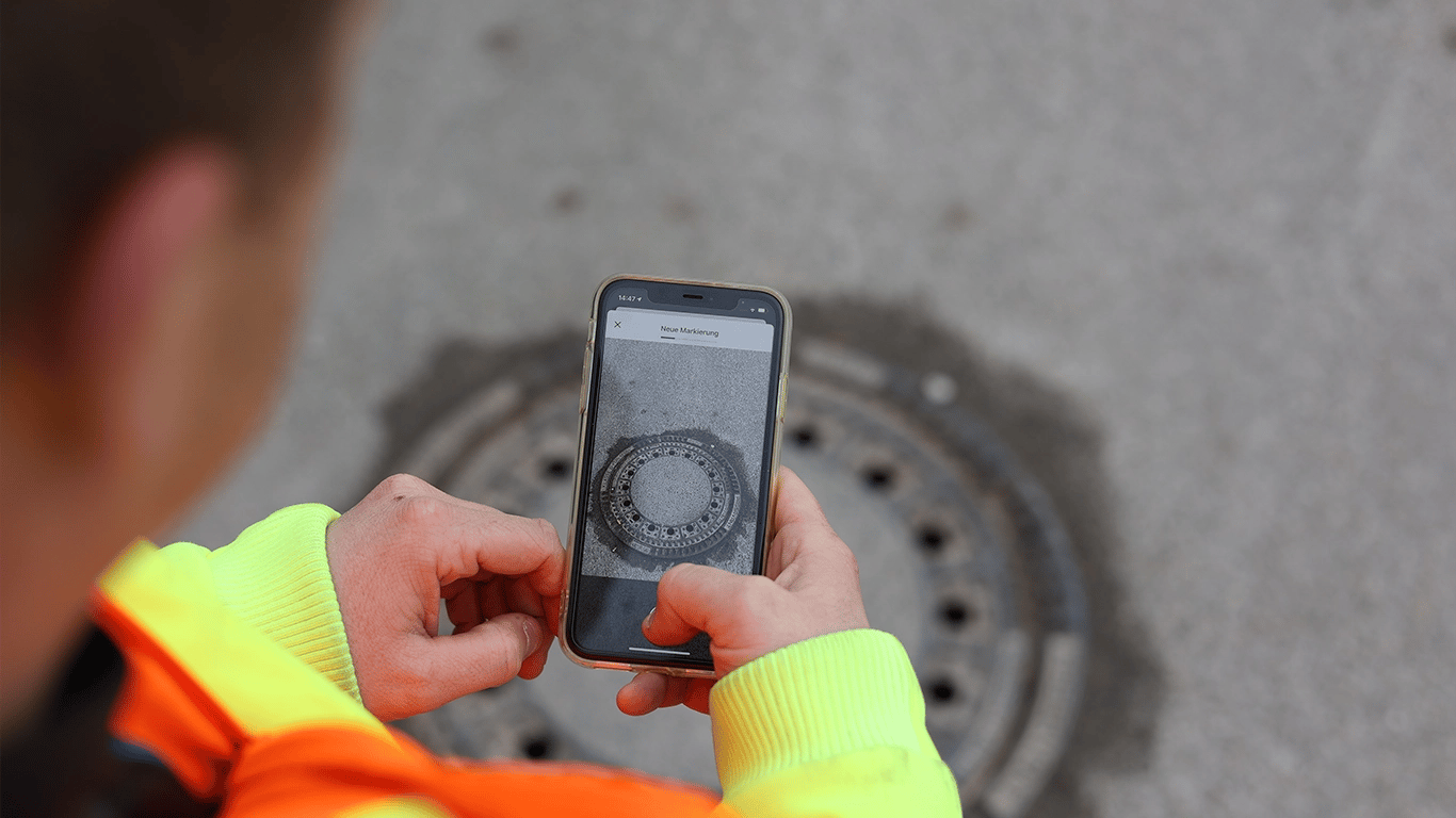 Ein Mitarbeiter des Bauhofs hält ein Handy in der Hand und hat das Markierungstool geöffnet. Im Hintergrund ist eine Schachtabdeckung zu sehen, eine Aufgabe der Verkehrssicherungspflicht